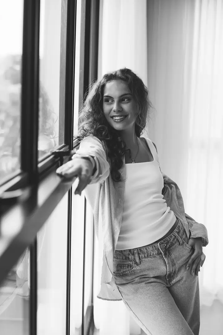 Mulher de camisa aberta e regata branca junto à janela, em preto e branco