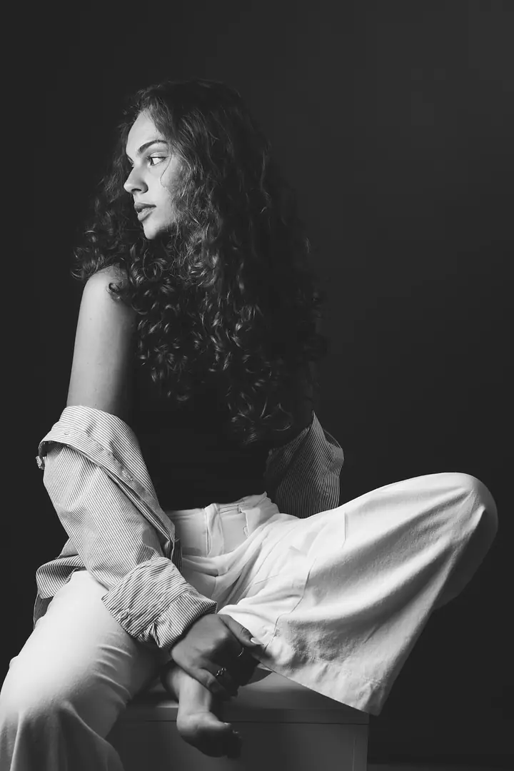 Mulher de cabelo cacheado sentada no chão, camisa e calça claras, preto e branco
