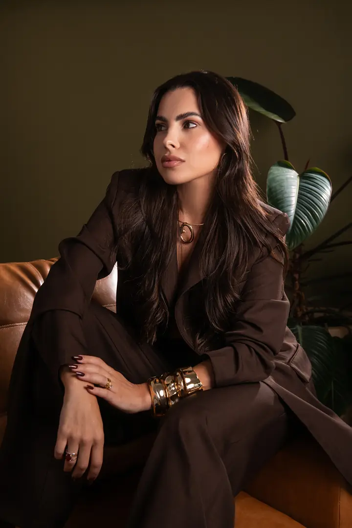 Mulher de conjunto marrom sentada em sofá, pulseiras douradas e planta ao fundo