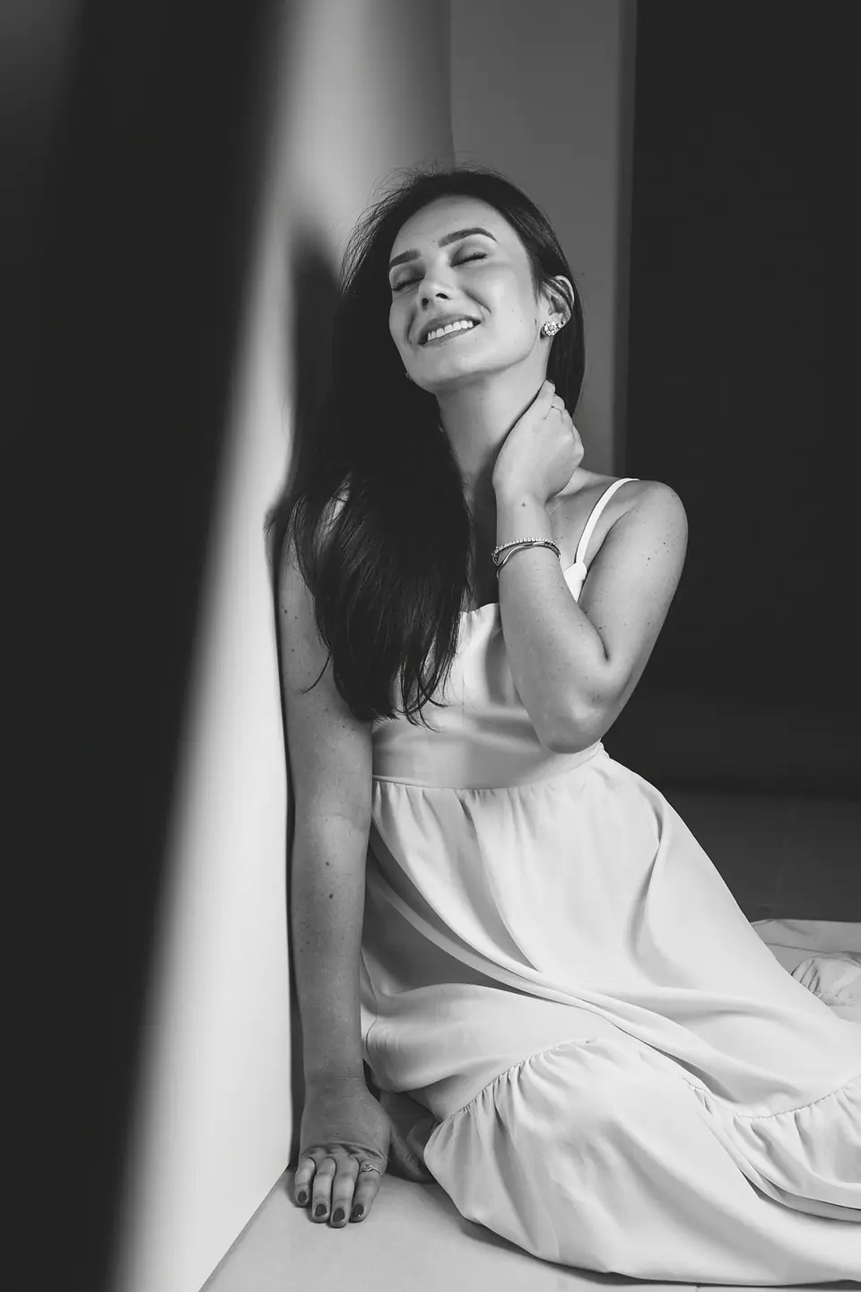 Mulher de vestido branco encostada na parede com a mão no pescoço, preto e branco