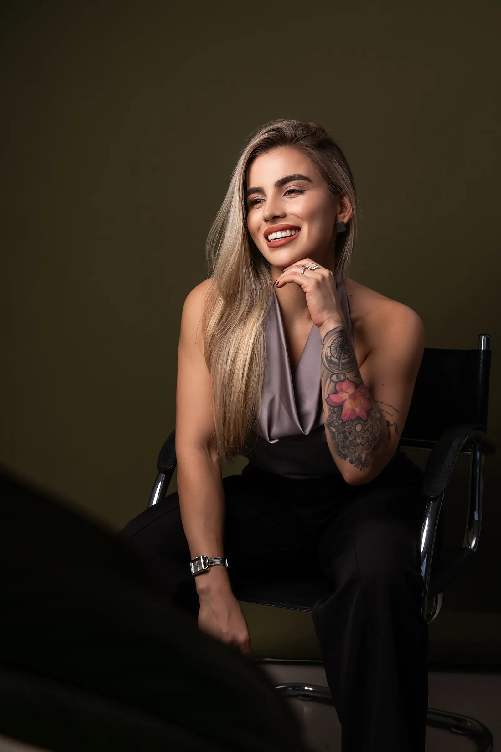 Mulher loira de blusa cinza sentada com a mão no queixo, fundo verde-escuro