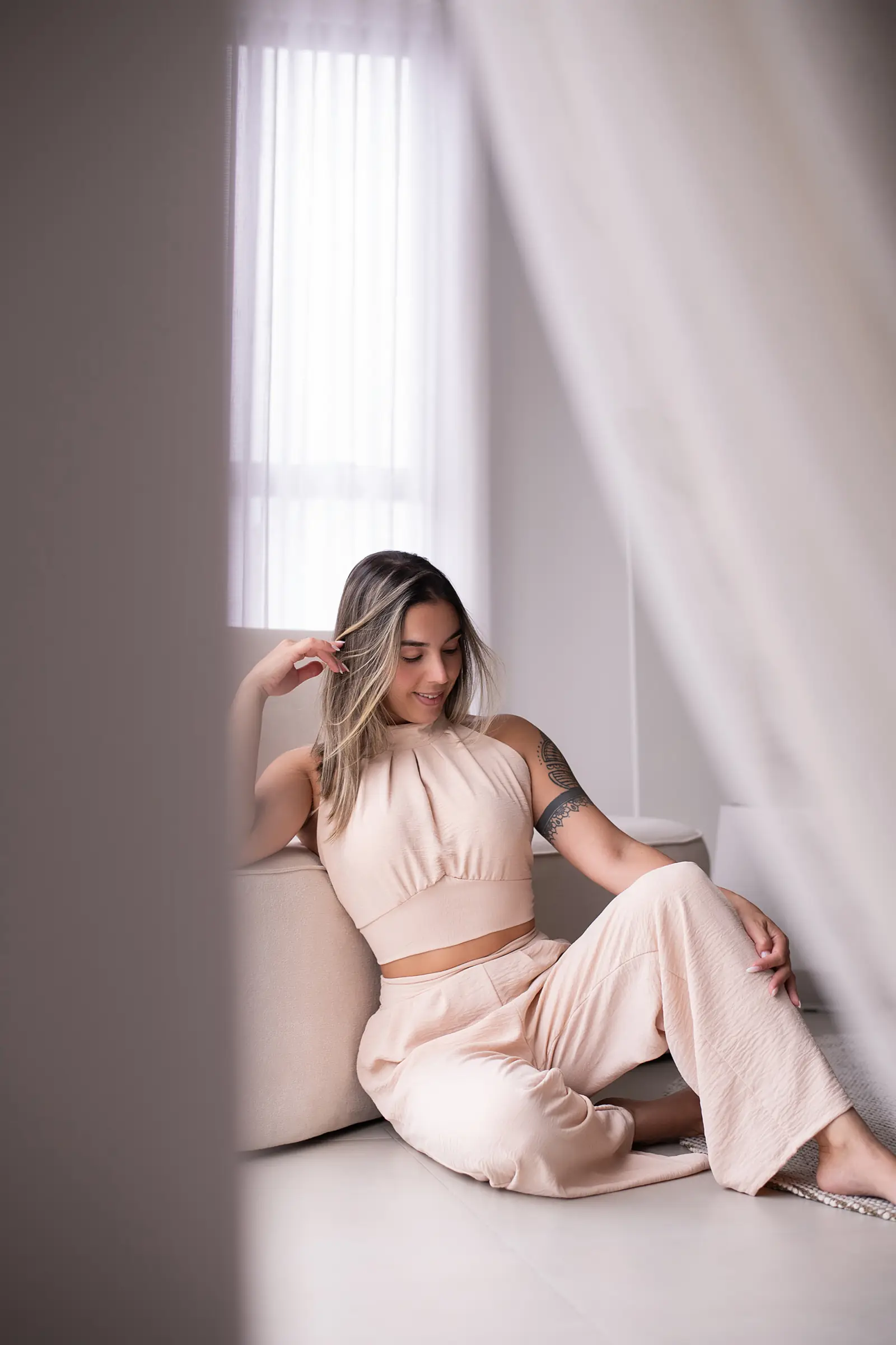 Mulher de conjunto rosa sentada junto à cortina, mãos no cabelo
