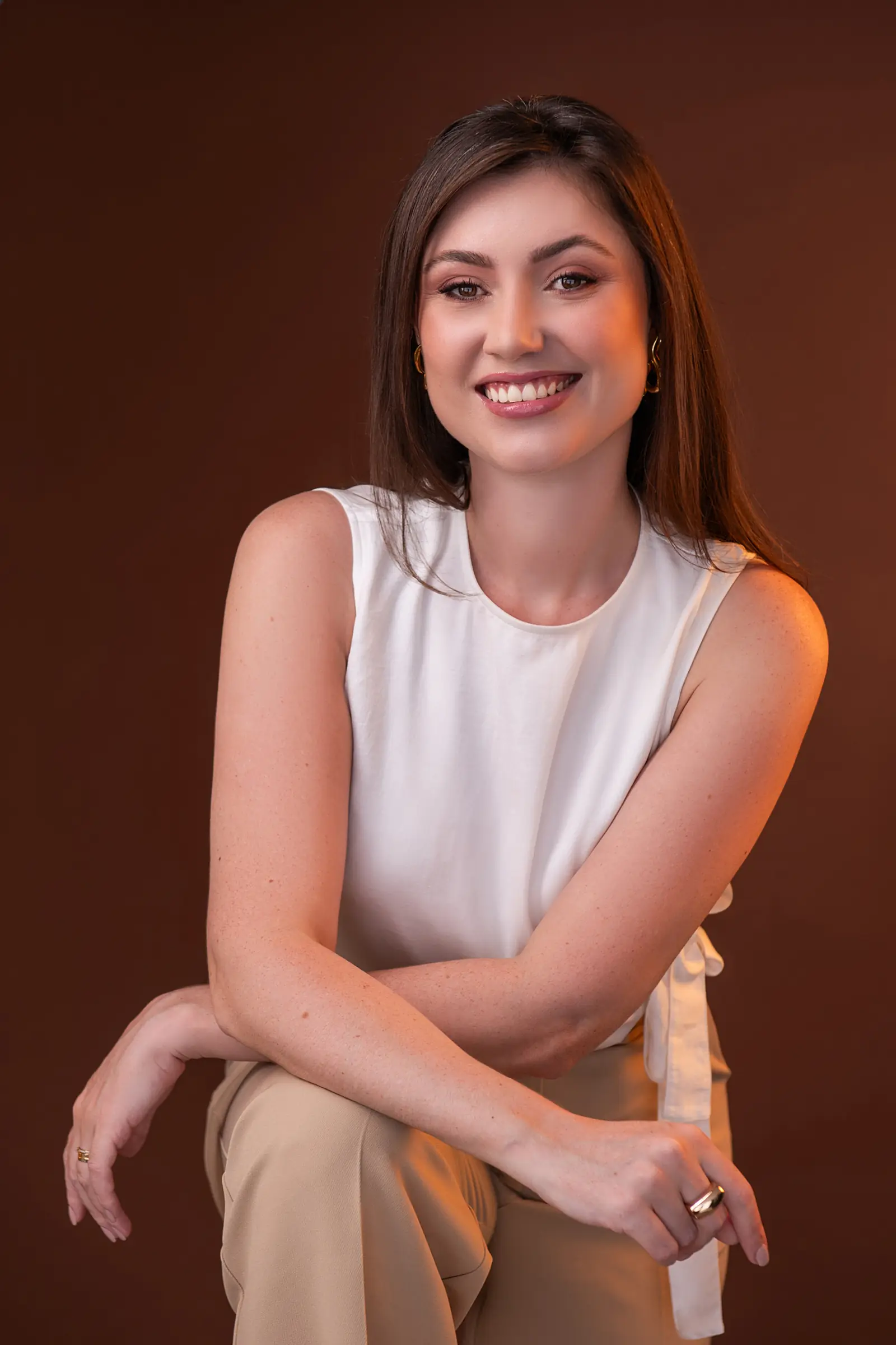 Mulher de blusa branca sem manga com os braços cruzados, sorrindo, fundo marrom