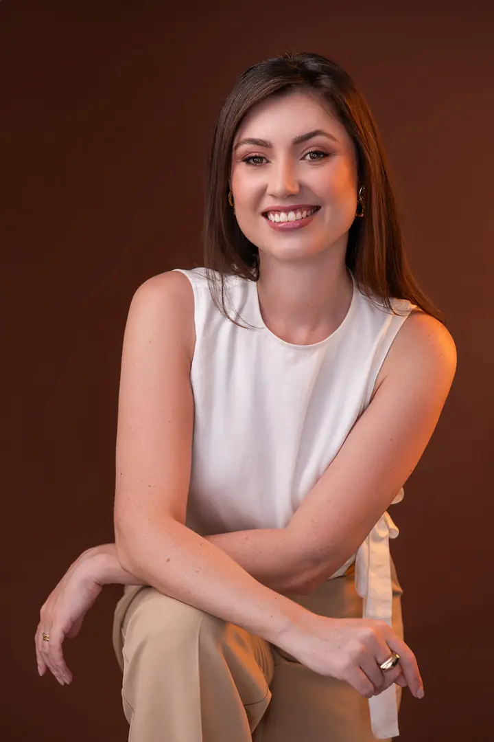 Mulher de blusa branca sem manga com os braços cruzados, sorrindo, fundo marrom