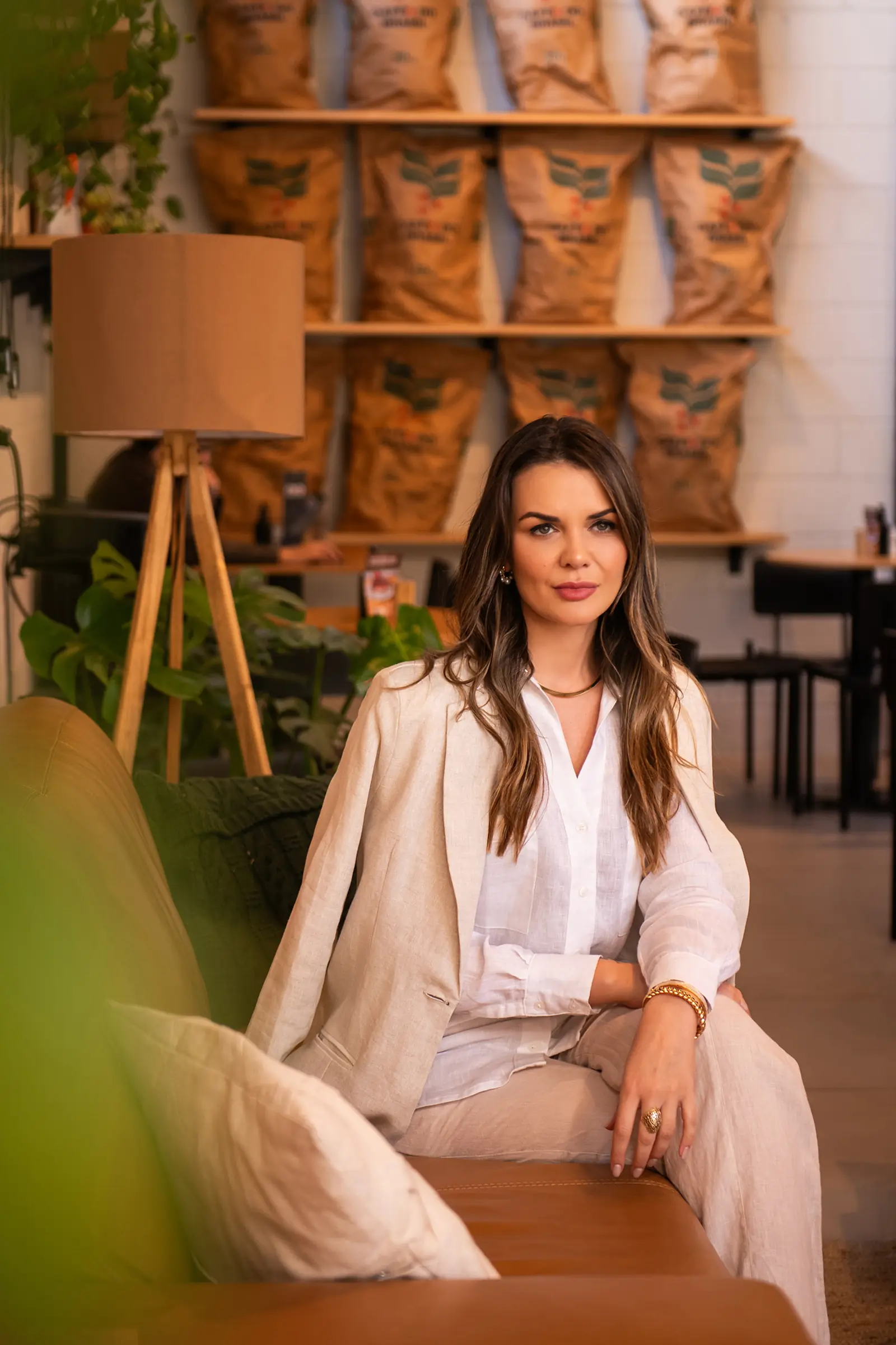 Mulher de blazer bege sentada em poltrona, ambiente de cafeteria ao fundo