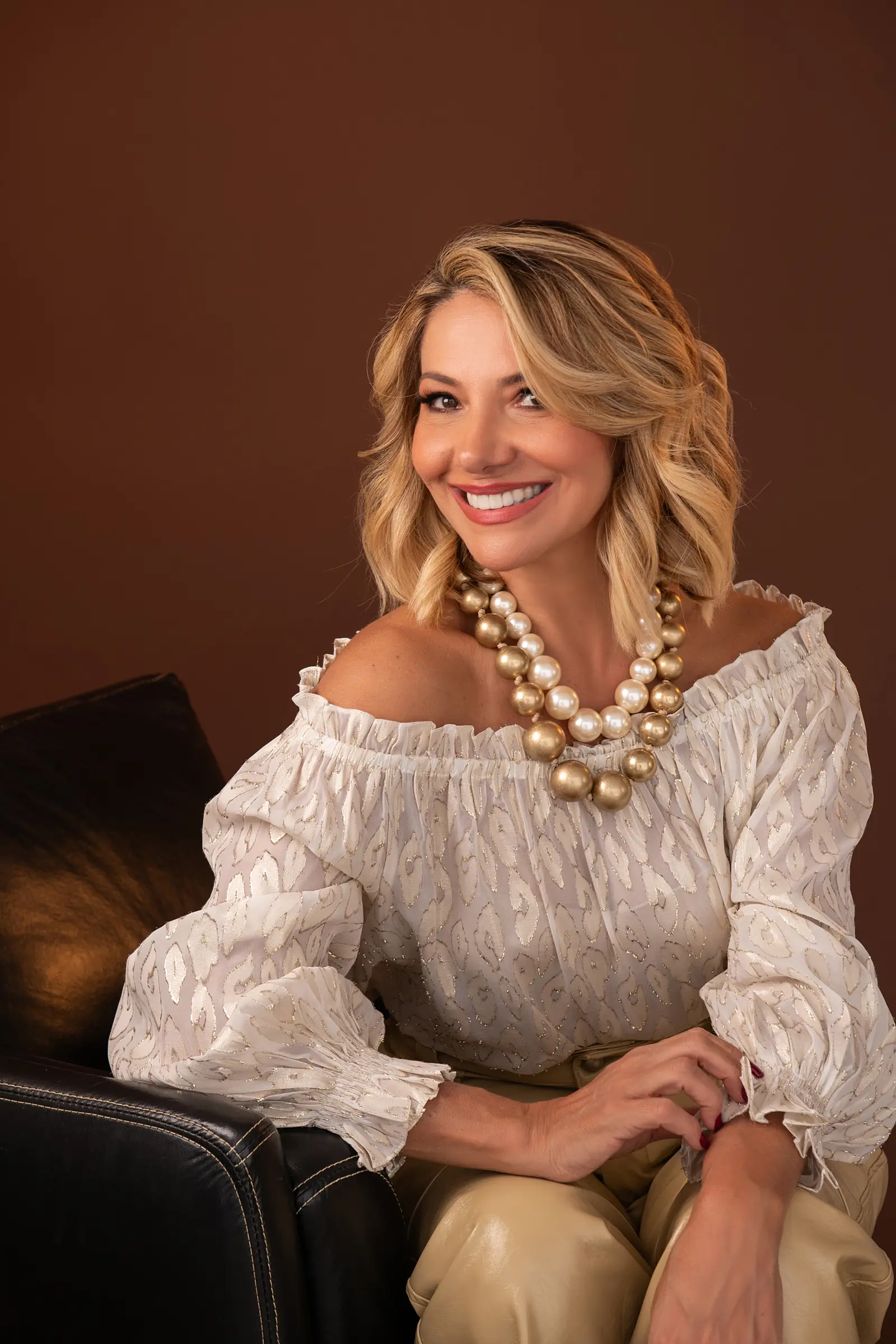 Mulher de blusa branca com colar de pérolas grandes, fundo marrom