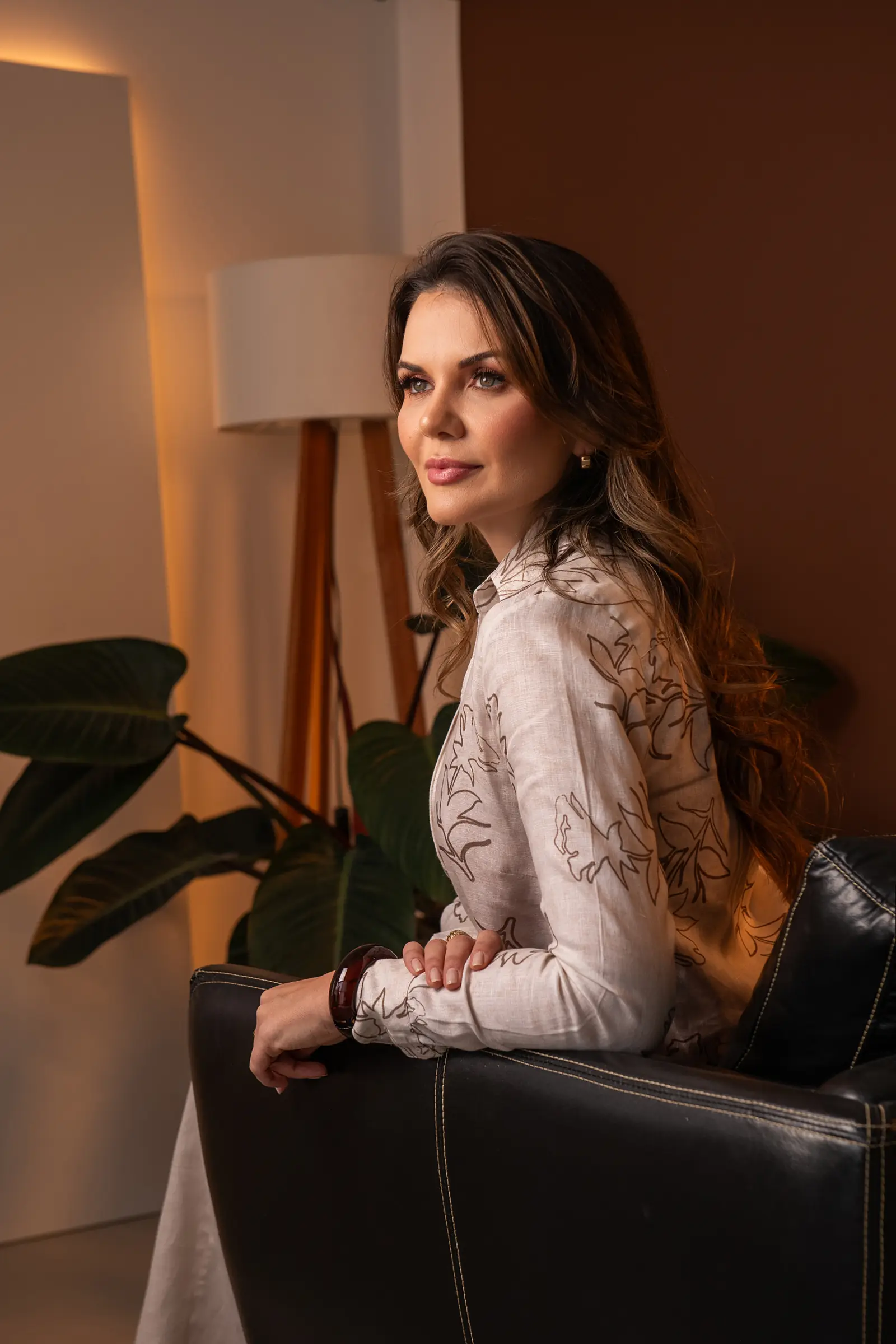 Mulher de blusa bordada sentada de lado, luminária e planta ao fundo