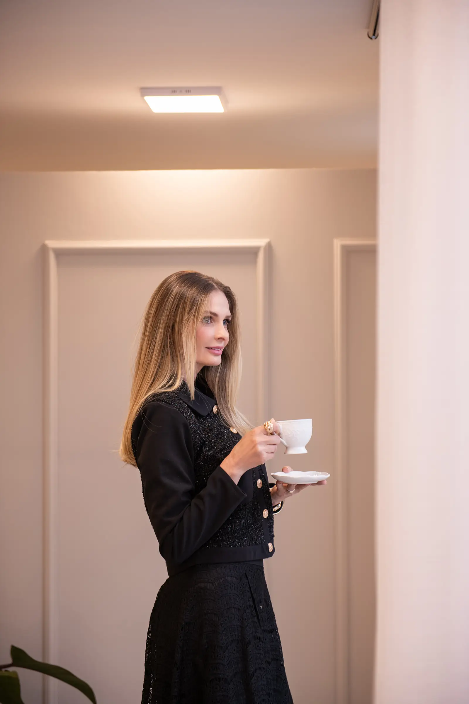 Mulher de blusa preta em pé segurando xícara de café, parede clara