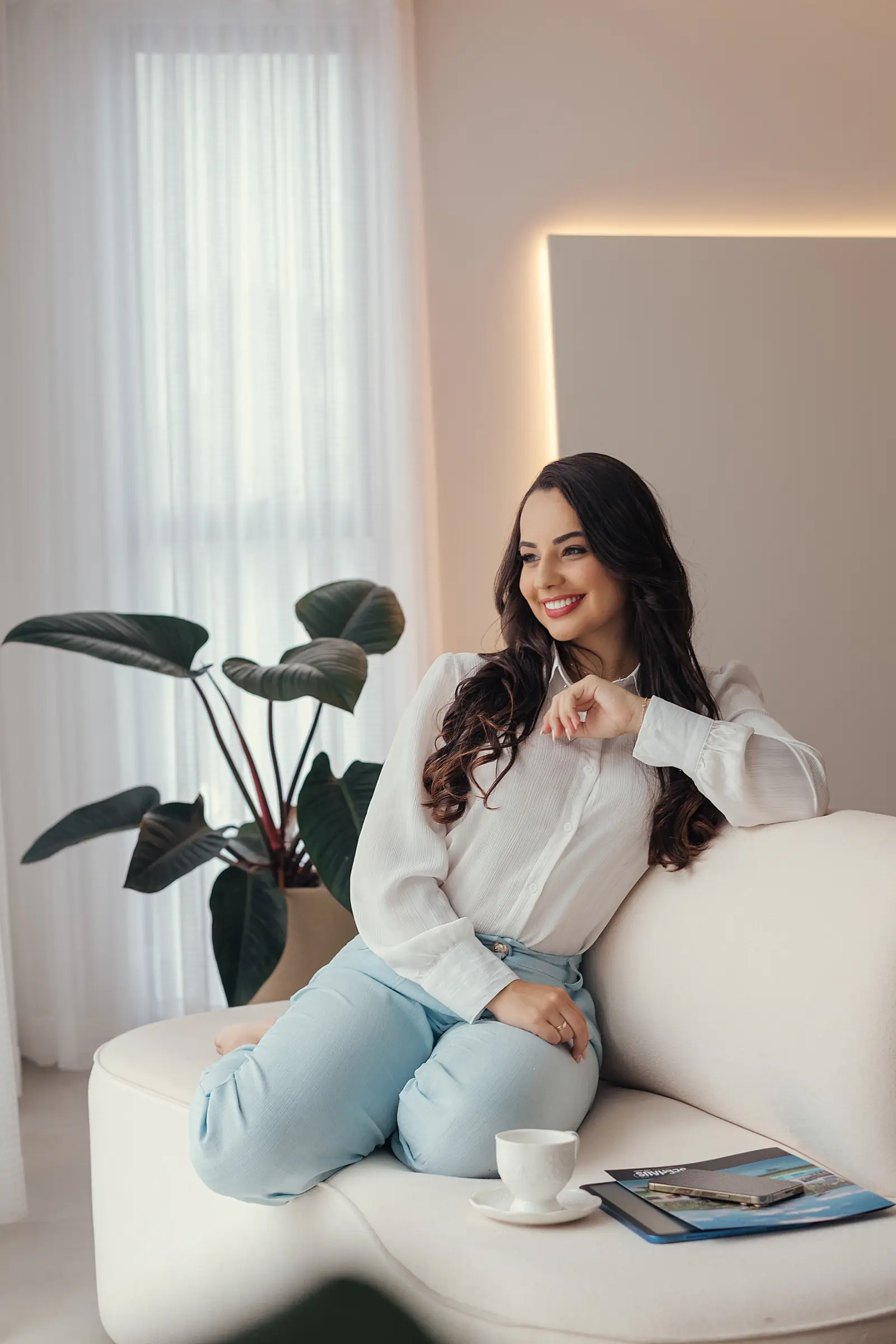 Mulher de blusa branca e calça jeans sentada em poltrona junto à cortina