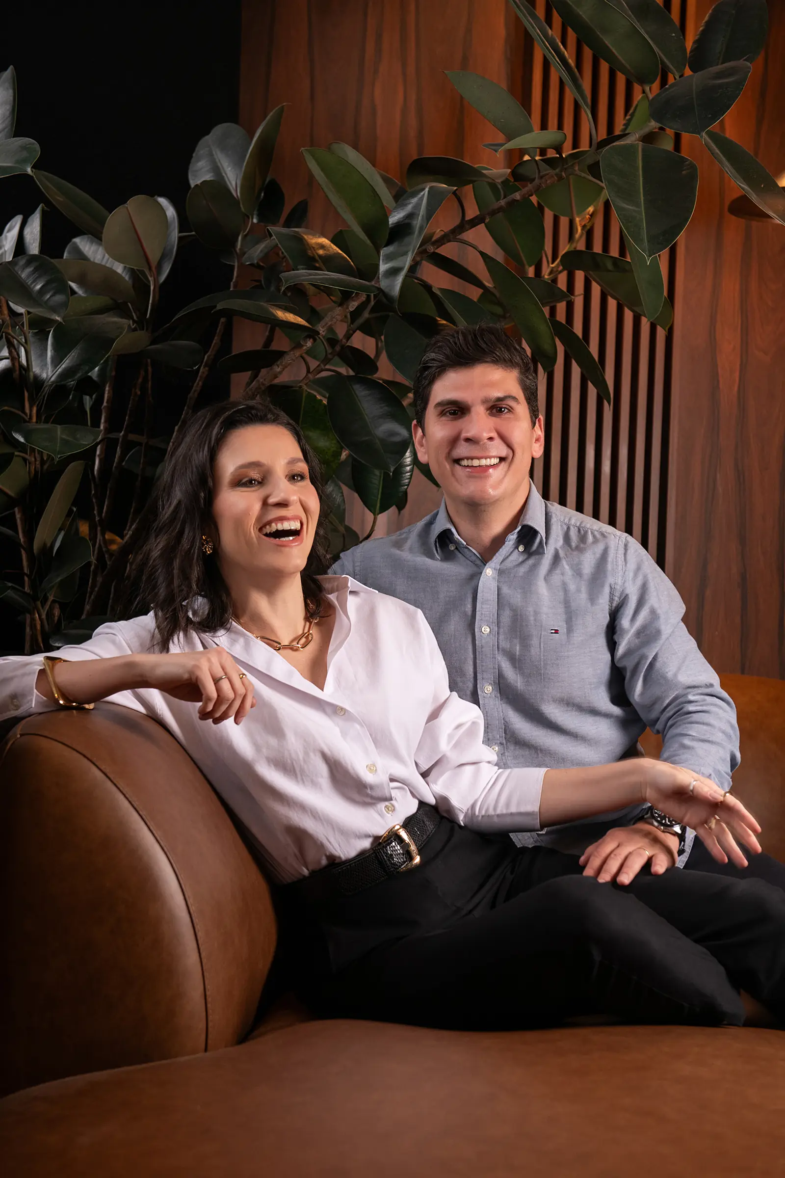 Casal de sócios rindo em sofá de couro, os dois de camisa clara