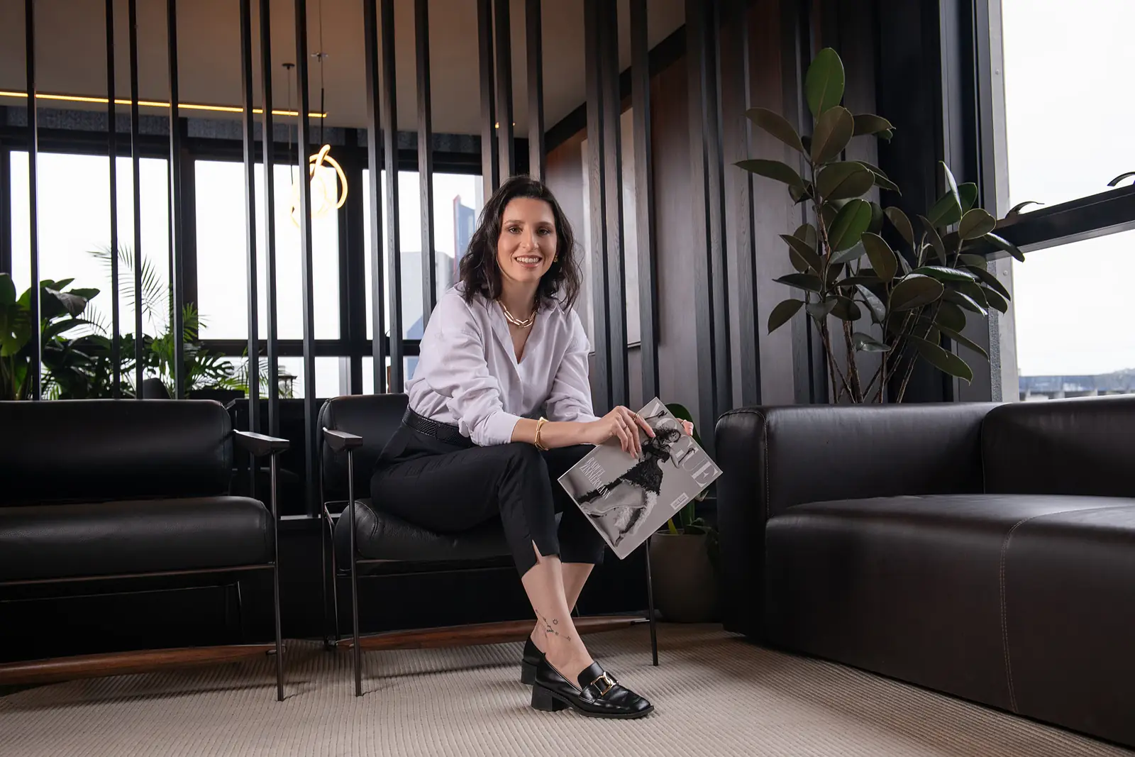 Mulher de camisa listrada sentada em sofá preto, escritório envidraçado