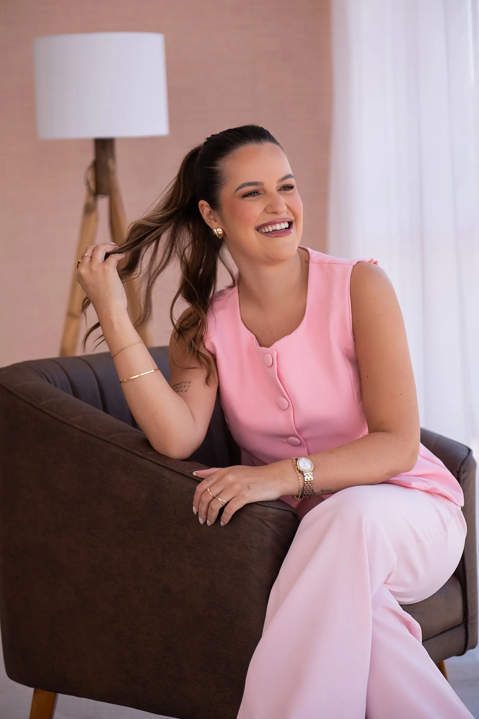 Mulher de blusa rosa sentada no braço do sofá, rindo, cortina clara atrás