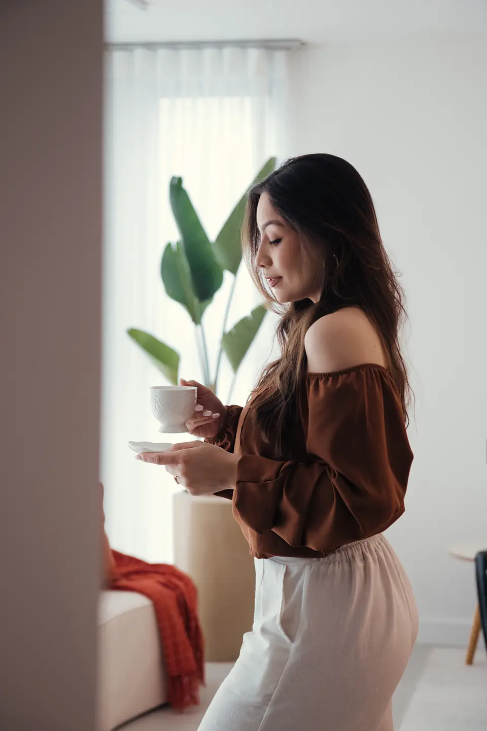 Mulher de blusa marrom ombro a ombro segurando xícara, planta ao lado