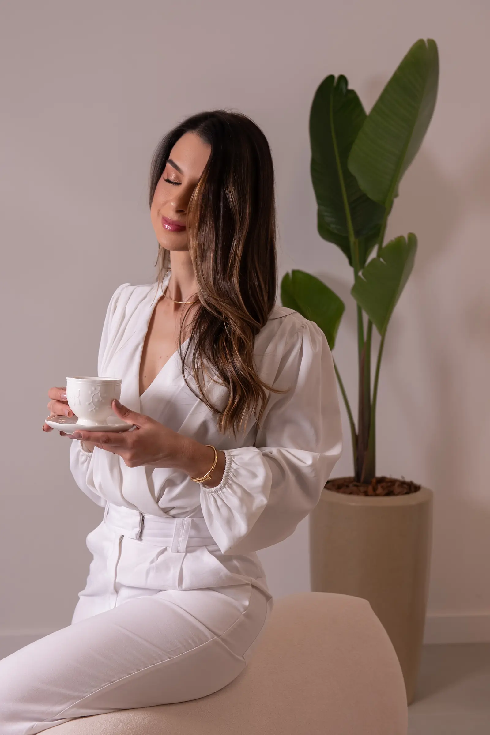 Mulher de conjunto branco segurando xícara de café, planta ao fundo