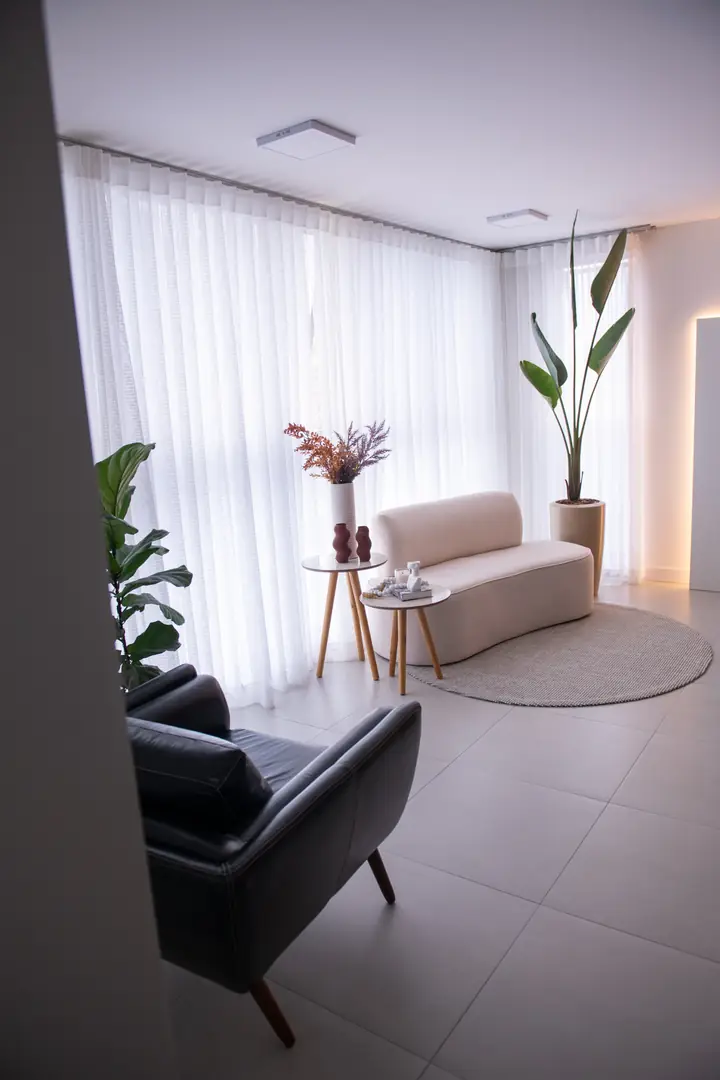 Vista ampla do estúdio com poltrona preta, sofá bege, tapete redondo e cortinas claras