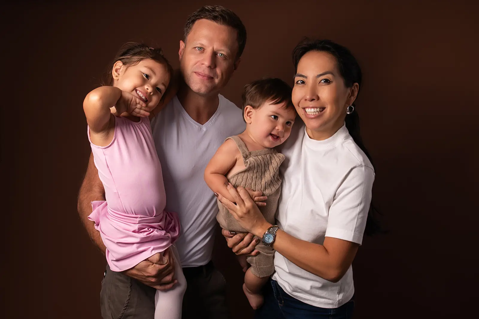 Família de quatro em pé, pais com a filha e o bebê no colo, fundo marrom