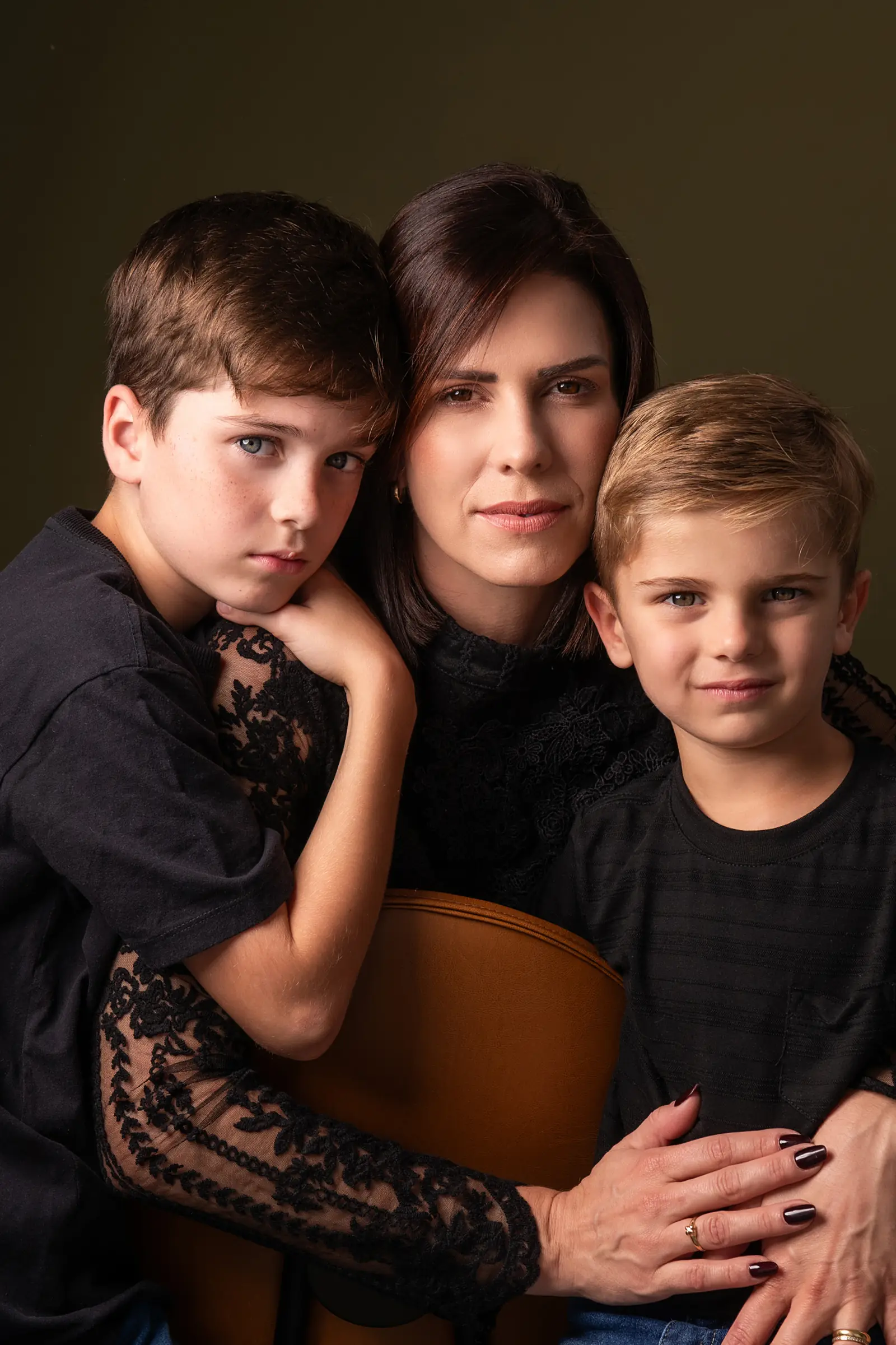 Mãe de blusa de renda entre os dois filhos meninos, fundo verde-escuro