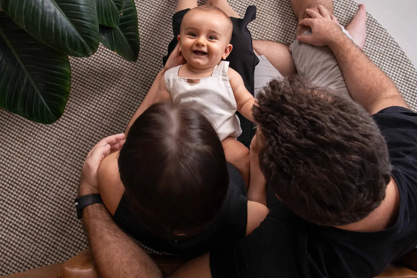 Pai segurando o bebê no alto enquanto a mãe deita no sofá, todos rindo