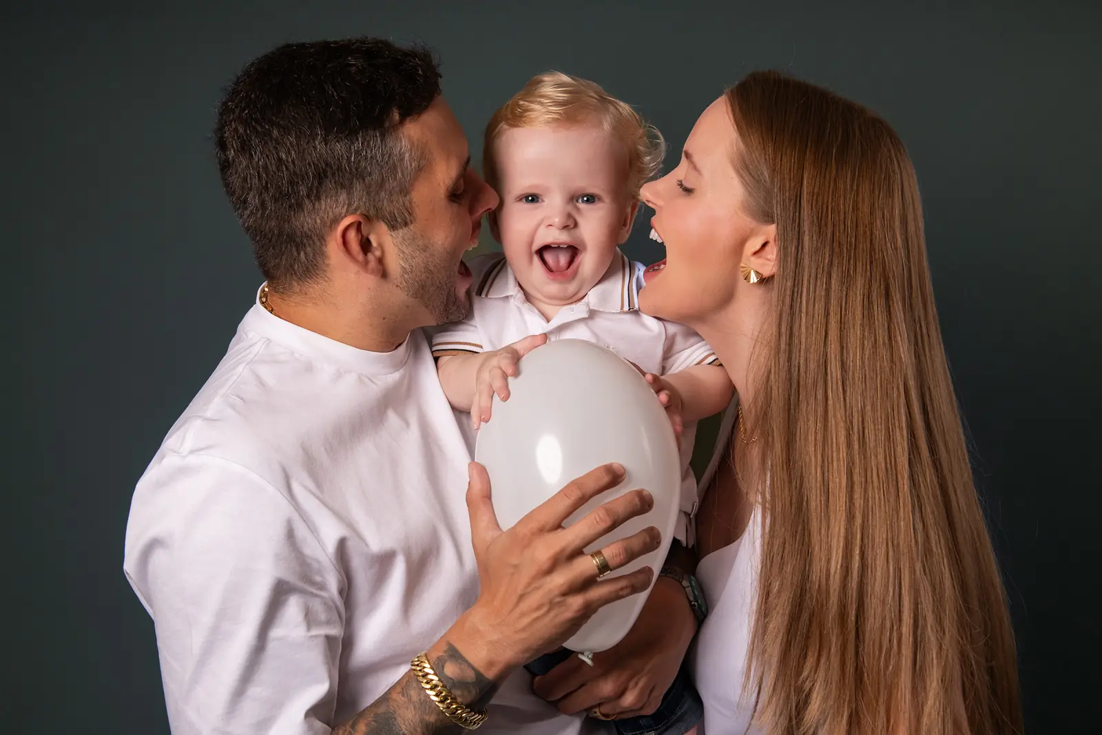 Pai e mãe beijando o filho pequeno que ri, fundo verde-escuro