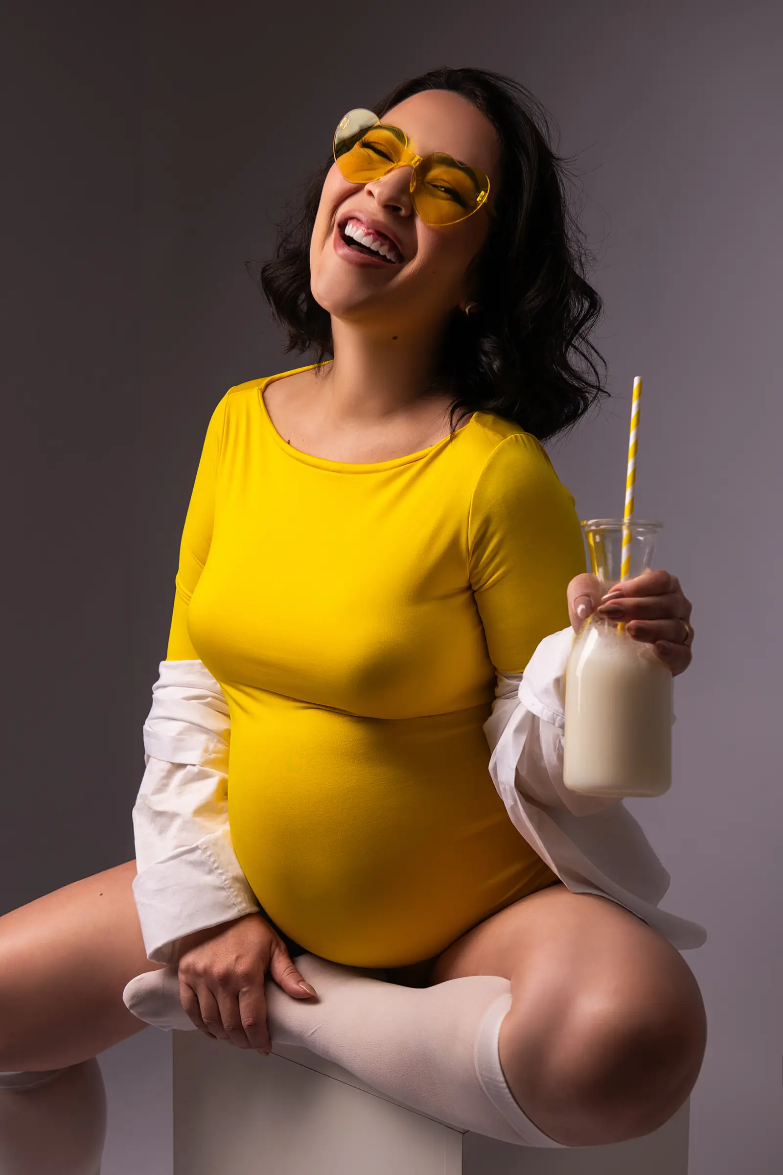 Gestante de body amarelo sentada rindo, camisa branca caída nos ombros