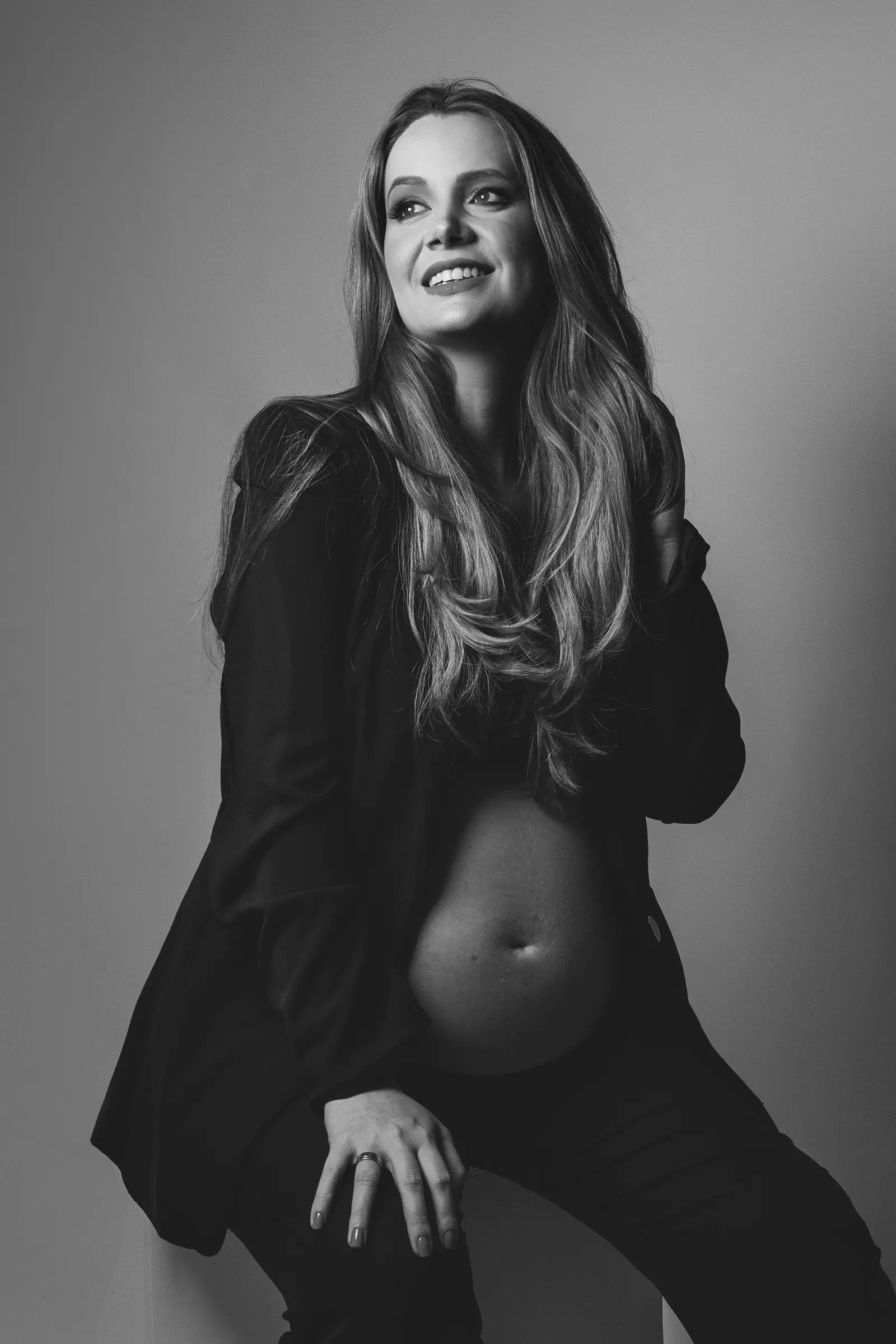 Gestante de blazer aberto sobre a barriga, sorrindo, preto e branco