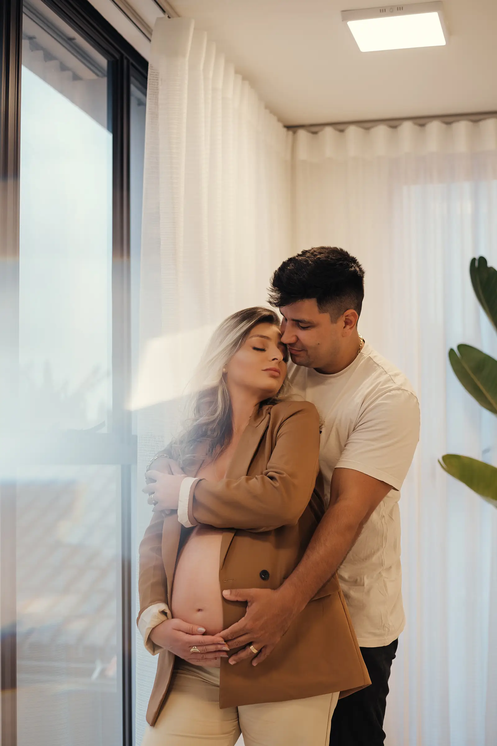Casal abraçado junto à janela, ele apoiado na barriga dela