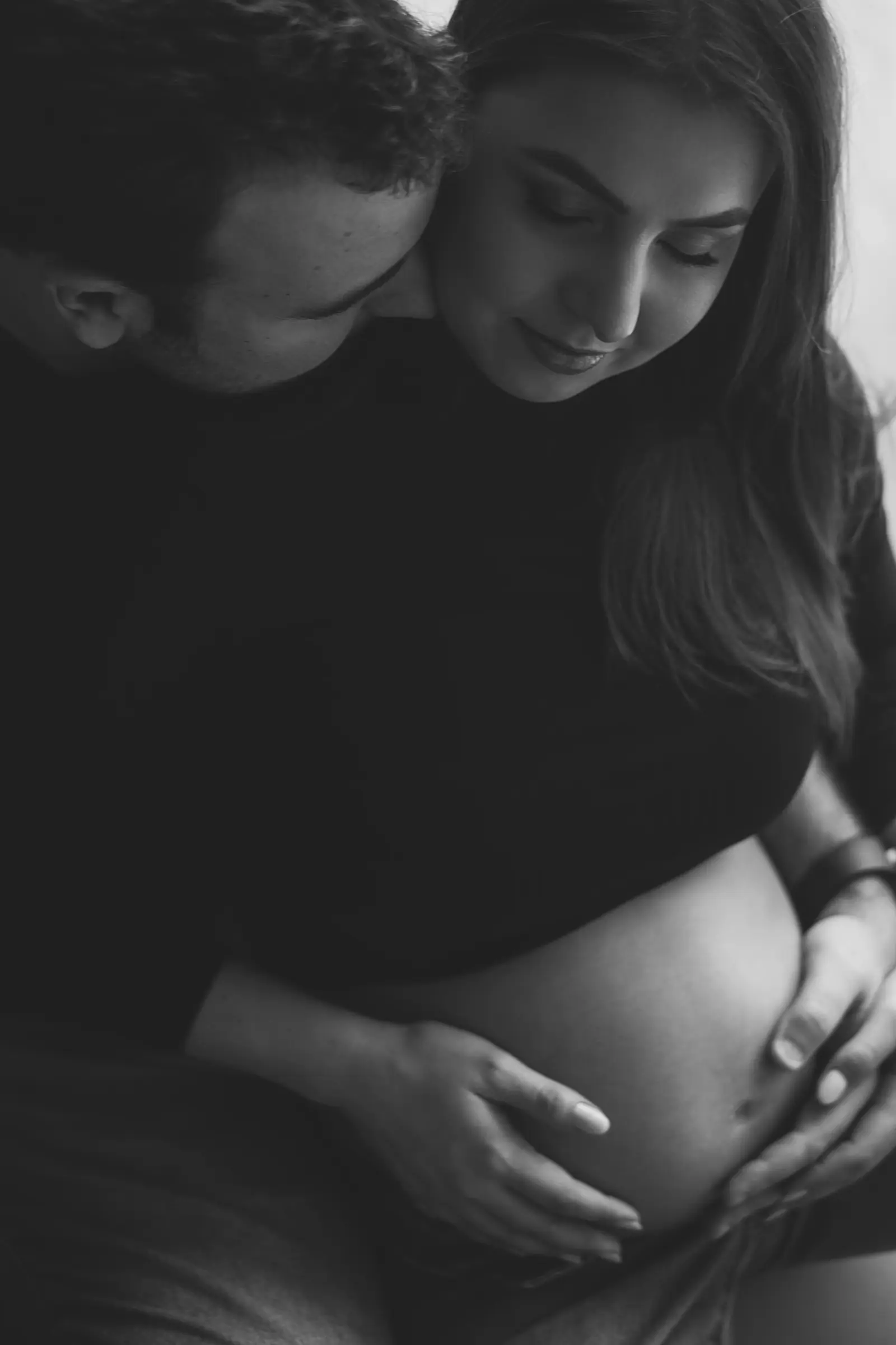 Mãos do casal sobre a barriga em luz baixa, preto e branco