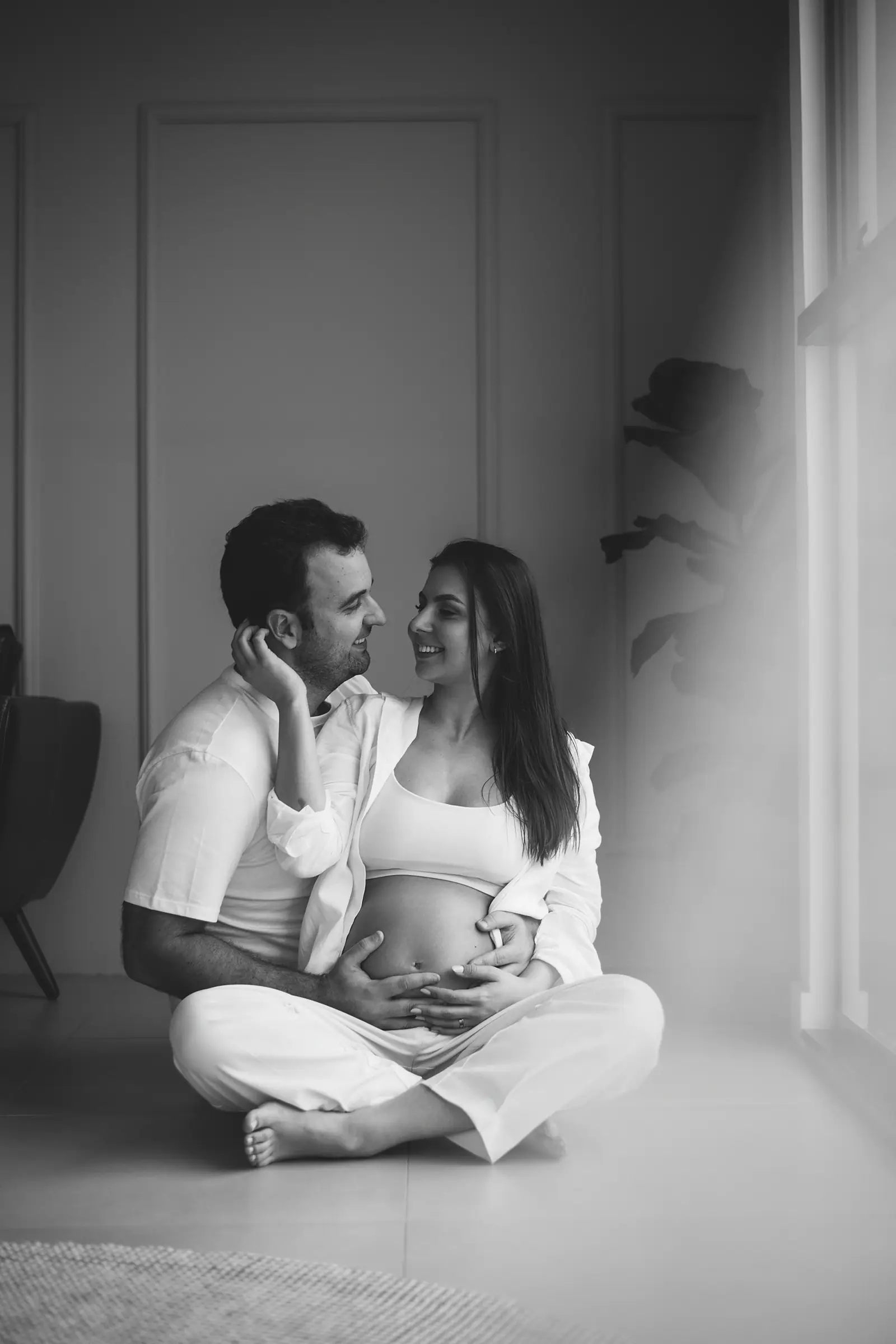 Casal sentado no chão de pernas cruzadas, mãos na barriga, preto e branco