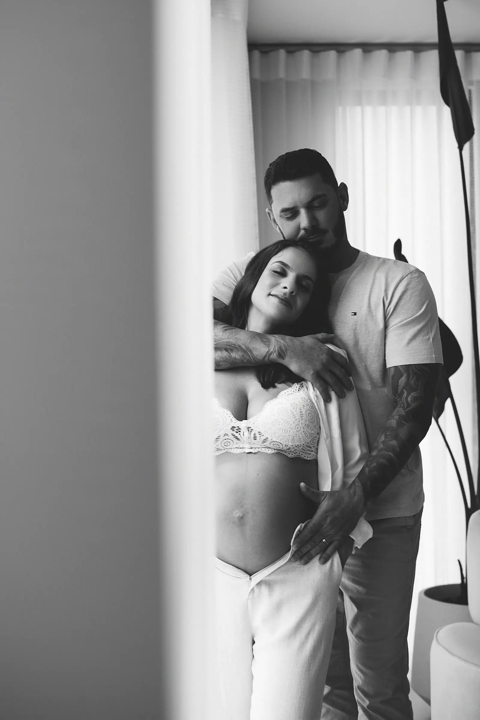 Casal abraçado de pé junto à cortina, mãos sobre a barriga