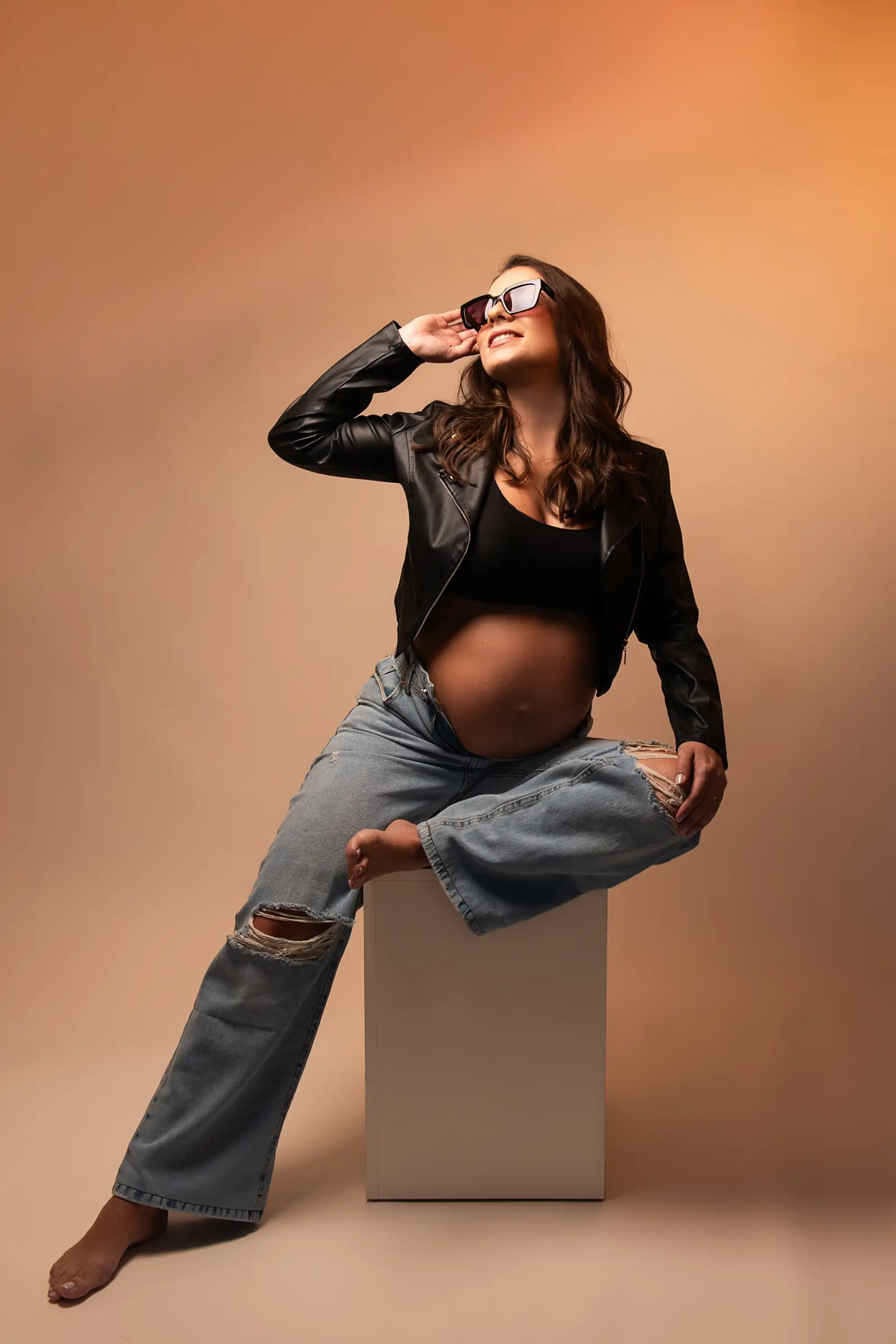 Gestante de jaqueta de couro e jeans sentada em cubo, fundo laranja